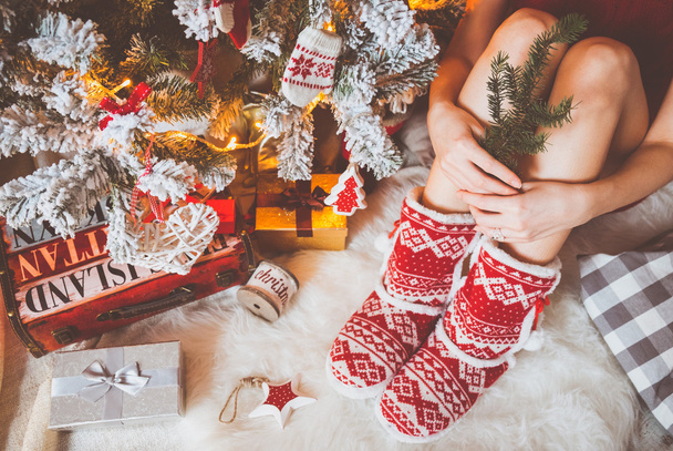 Young pretty woman in warm cozy clothes is sitting on the light wooden floor in her bright house near the christmas tree and holds gift box in her hands. - Фото, изображение