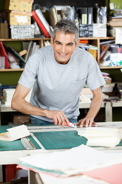 Happy Mature Male Worker Cutting Paper At Table - Valokuva, kuva