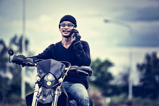 Aasia komea mies moottoripyöräilijä soittaa puhelimeen moottoripyörällä
 - Valokuva, kuva