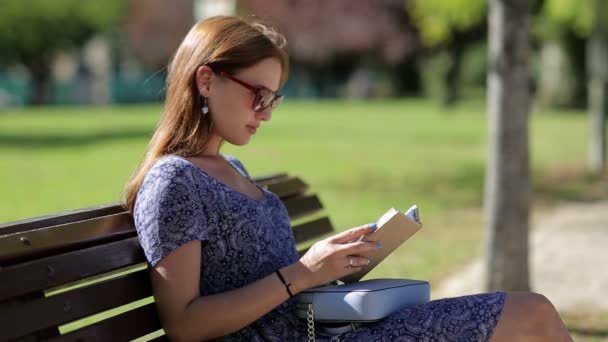 Young woman reading a book and sitting on a bench outside in a park in summer, in sunglasses with handbag - Πλάνα, βίντεο