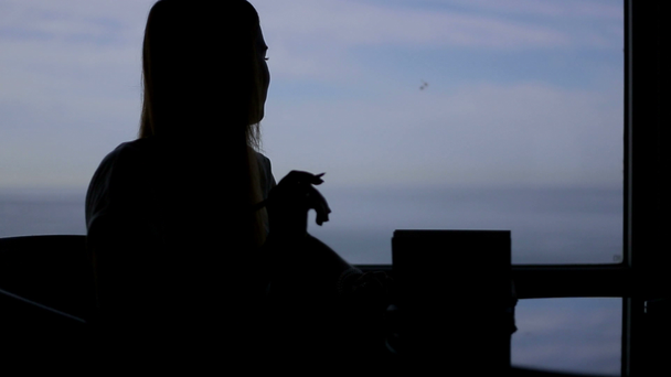 silhouette of young beautiful girl sits in a cafe with panoramic window sea view . Morning, with a book, notepad, wrighting, thinking - Footage, Video