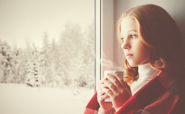 nachdenklich trauriges Mädchen mit einem wärmenden Getränk schaut aus dem Fenster in - Foto, Bild