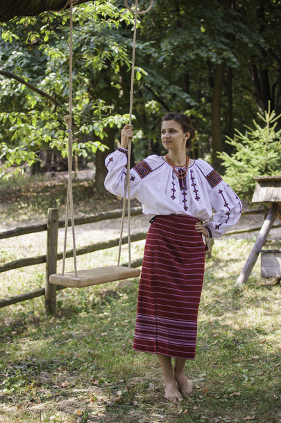 Beautiful girl in Ukrainian national dress stands near the swing - Photo, Image