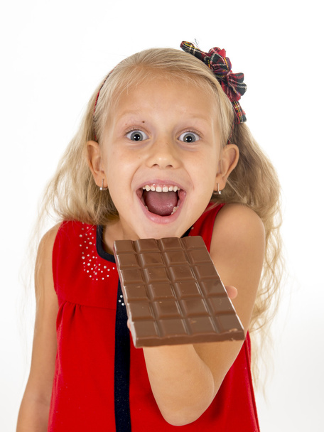 little beautiful female child in red dress holding happy delicious chocolate bar in her hands eating delighted - Photo, image