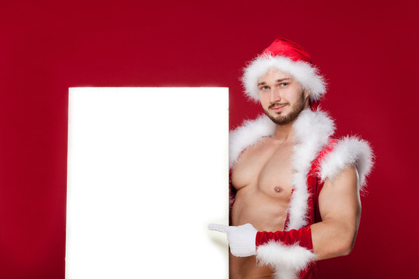the very muscular  bronzed handsome sexy Santa Claus on red  background, hold   placard and smile, isolated - Photo, image