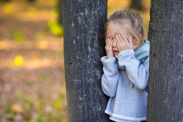 小さな女の子が遊んでかくれんぼ秋の公園で木の近く - 写真・画像