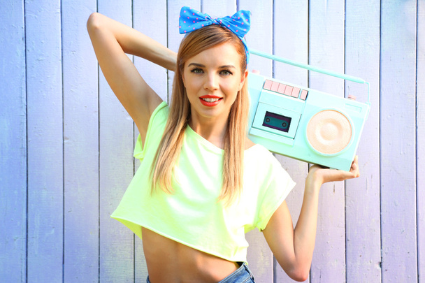 Beautiful girl with pretty smile holding retro tape recorder on color wooden background - Fotoğraf, Görsel