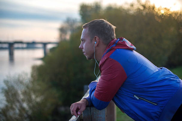 Attractive young athletic man standing on the beach and looks into distance of  river to  bridge, listening music - Фото, зображення