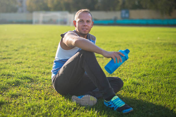 Sporting an attractive man sitting on grass and rests in the stadium, keeps shaker, sunny day - Фото, зображення
