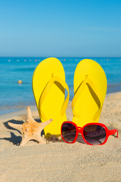 Travel by sea. Beach vacation. Red sunglasses and yellow flip-flops on sandy seashore. - Zdjęcie, obraz
