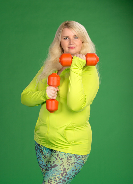 Plus size woman pumping up muscules with dumbbells on green back - Fotó, kép