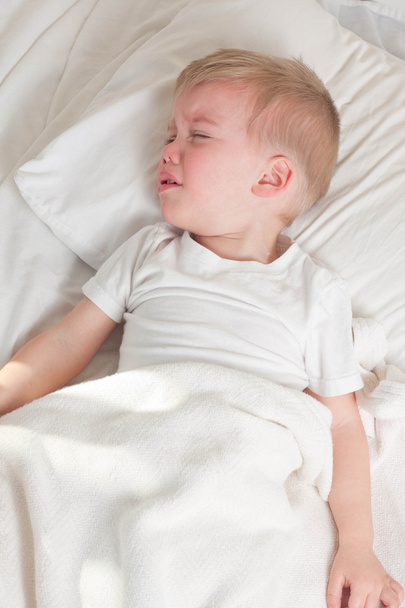 ein krankes blondes Kleinkind mit weißem T-Shirt liegt im Bett - Foto, Bild