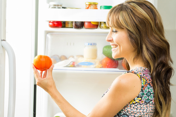 Femme portant une robe colorée dans la cuisine moderne ouvrant la porte du réfrigérateur et tenant un shile de tomate souriant, angle de profil
 - Photo, image