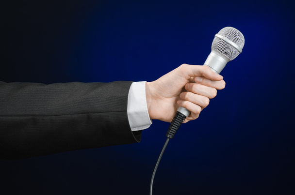Business and speech topic: Man in black suit holding a microphone on a gray dark blue isolated background in studio - Фото, изображение