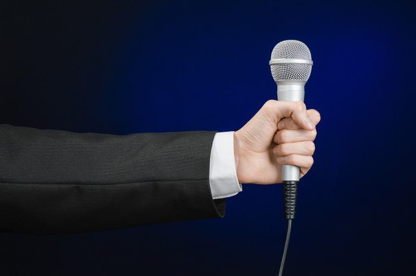 Business and speech topic: Man in black suit holding a microphone on a gray dark blue isolated background in studio - Foto, afbeelding