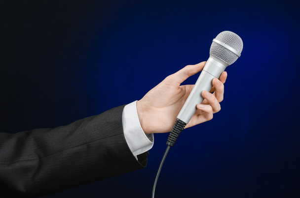 Business and speech topic: Man in black suit holding a microphone on a gray dark blue isolated background in studio - Fotografie, Obrázek