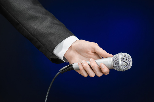 Business and speech topic: Man in black suit holding a microphone on a gray dark blue isolated background in studio - Foto, Bild