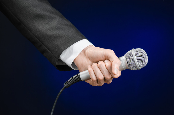 Business and speech topic: Man in black suit holding a microphone on a gray dark blue isolated background in studio - Foto, immagini