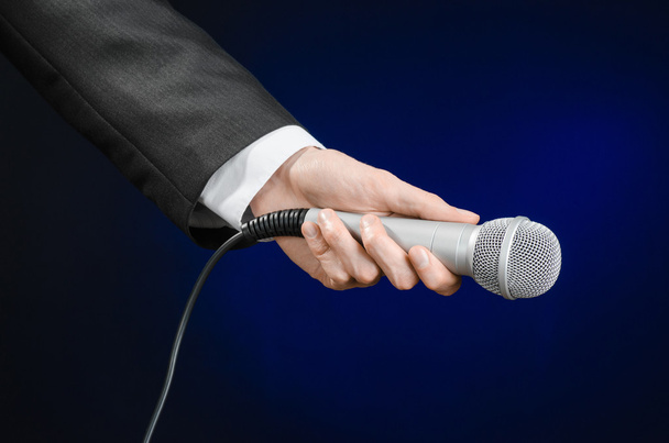 Business and speech topic: Man in black suit holding a microphone on a gray dark blue isolated background in studio - Foto, afbeelding