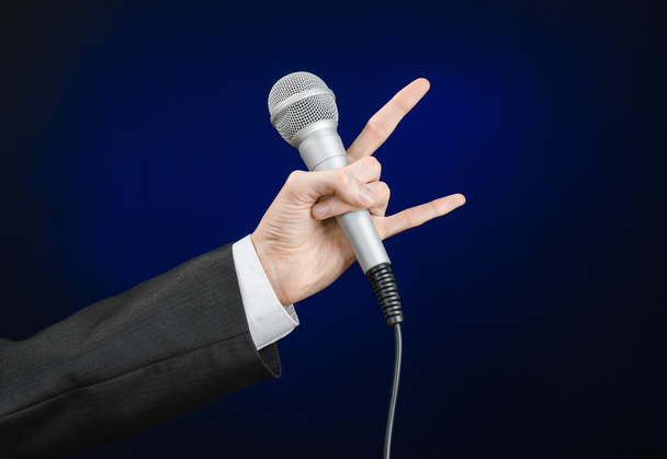 Business and speech topic: Man in black suit holding a microphone on a gray dark blue isolated background in studio - Fotó, kép