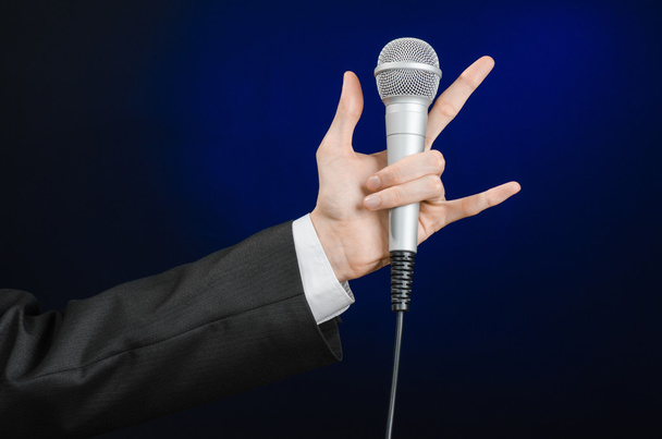 Business and speech topic: Man in black suit holding a microphone on a gray dark blue isolated background in studio - Fotografie, Obrázek