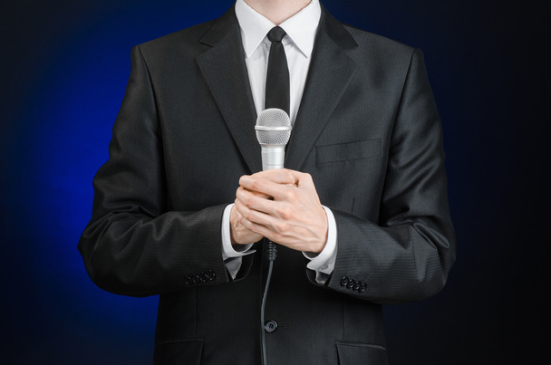 Business and speech topic: Man in black suit holding a microphone on a gray dark blue isolated background in studio - Zdjęcie, obraz