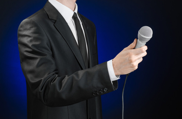 Business and speech topic: Man in black suit holding a microphone on a gray dark blue isolated background in studio - Fotó, kép