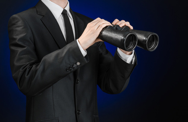 Business and search topic: Man in black suit holding a black binoculars in hand on a dark blue background in studio isolated - 写真・画像