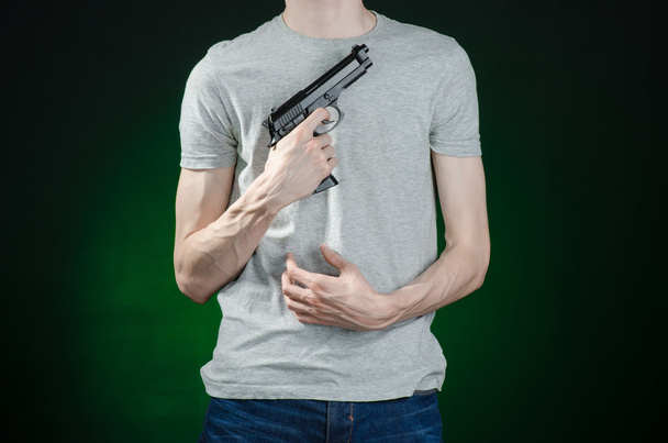 Firearms and murderer topic: man in a gray t-shirt holding a gun on a dark green background isolated in studio - Photo, image