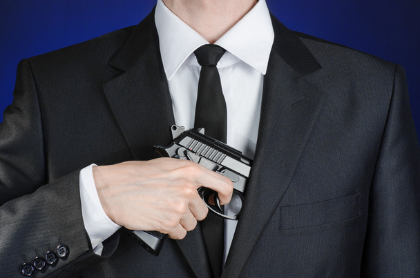 Firearms and security topic: a man in a black suit holding a gun on a dark blue background in studio isolated - Φωτογραφία, εικόνα