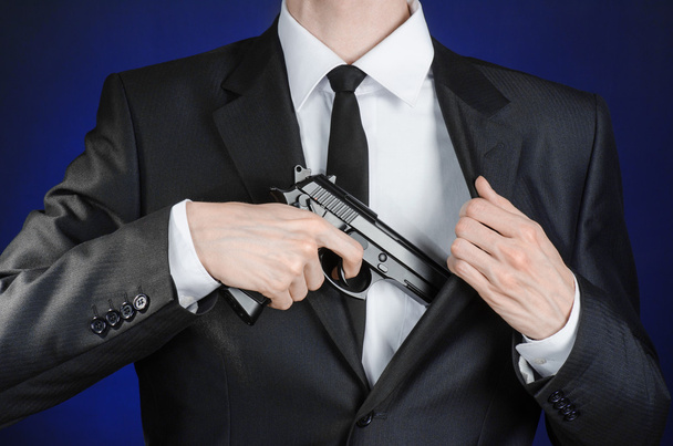 Armas de fuego y temas de seguridad: un hombre con un traje negro sosteniendo un arma sobre un fondo azul oscuro en un estudio aislado
 - Foto, imagen