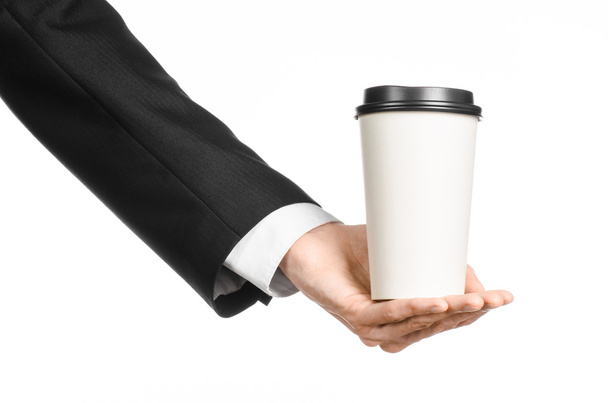 Business lunches coffee theme: businessman in a black suit holding a white blank paper cup of coffee with a brown plastic cap isolated on a white background in the studio, advertising coffee - Foto, Imagen