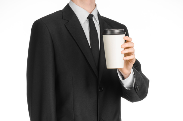 Business lunches coffee theme: businessman in a black suit holding a white blank paper cup of coffee with a brown plastic cap isolated on a white background in the studio, advertising coffee - Foto, immagini