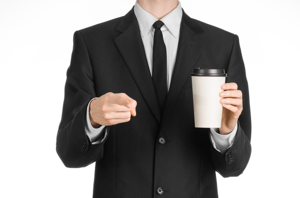 Business lunches coffee theme: businessman in a black suit holding a white blank paper cup of coffee with a brown plastic cap isolated on a white background in the studio, advertising coffee - Foto, afbeelding