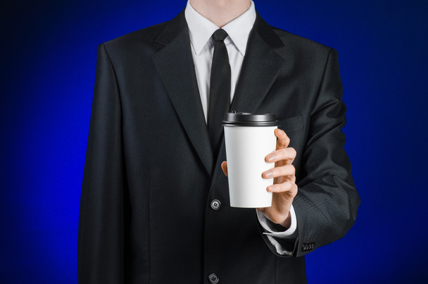 Business lunch and coffee theme: businessman in a black suit holding a white blank paper cup of coffee with a brown plastic cap on a dark blue background isolated in the studio, advertising coffee - Фото, изображение