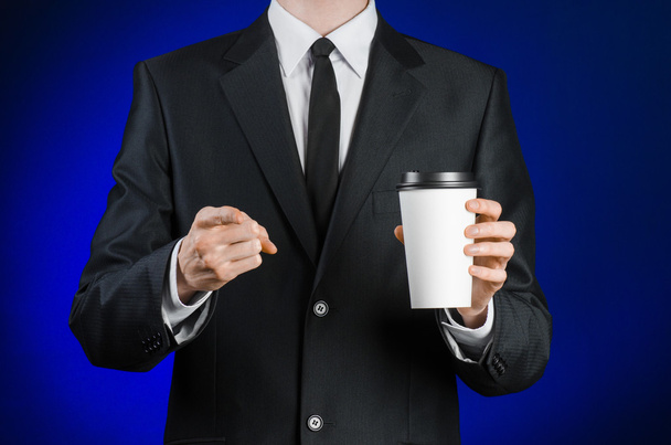 Business lunch and coffee theme: businessman in a black suit holding a white blank paper cup of coffee with a brown plastic cap on a dark blue background isolated in the studio, advertising coffee - Foto, Imagem