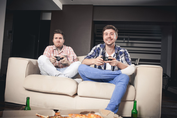Two young men playing video games while sitting on sofa - Foto, afbeelding