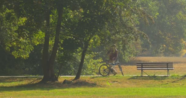 男のバックパックは、ベンチの男に立って戻って半ばショット自転車は立っている男の自転車になっているとの距離を乗って、公園への道で - 映像、動画