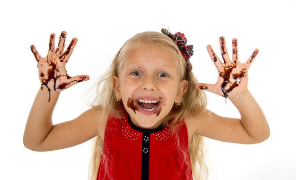pretty little female child with long blond hair and blue eyes wearing red dress showing dirty hands with stains of chocolate syrup - Photo, Image
