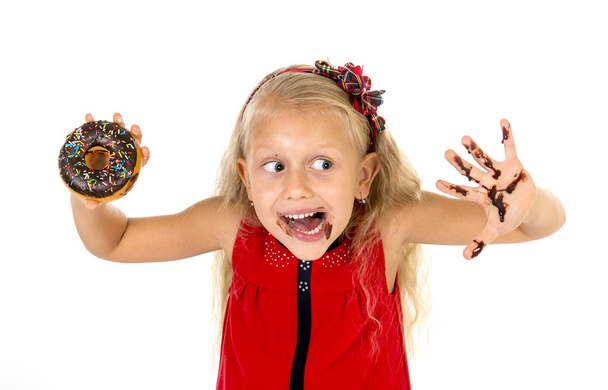 beautiful female child with blue eyes in cute red dress eating chocolate donut with syrup stains - Foto, afbeelding
