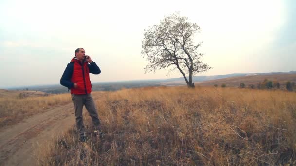 man reiziger praten aan de telefoon aard eenzame boom in de herfst in een rood jasje-reizen - Video