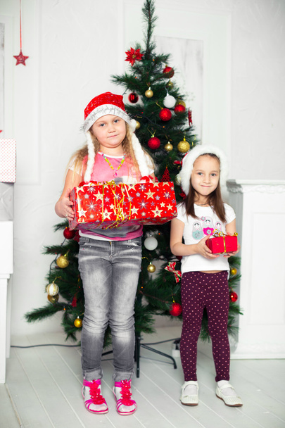 Two girls in Christmas costumes stand beside the tree with gifts their hands - Fotó, kép