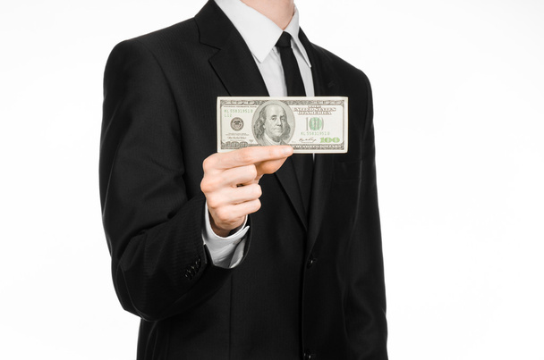Money and business theme: a man in a black suit holding a bill of 100 dollars and features a hand gesture on an isolated white background in studio - Zdjęcie, obraz