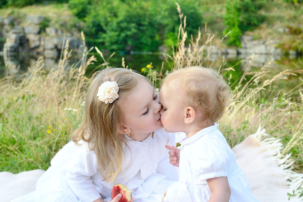 Дві маленькі сестри, що сидять на відкритому повітрі, цілуються один з одним
 - Фото, зображення
