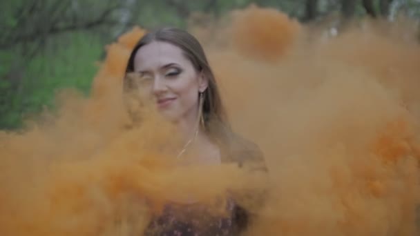 Happy young woman in dark dress with floral print having fun outdoor in summer forest. Beauty girl holding a colorful orange smoke bomb and goes to the camera.wonderful smile. medium close up slow - Footage, Video