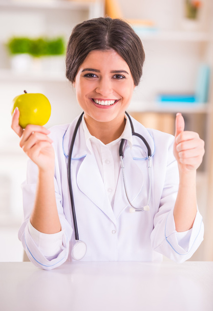 Portrait of a young beautiful woman doctor in white coat holding an apple in his office - Photo, Image