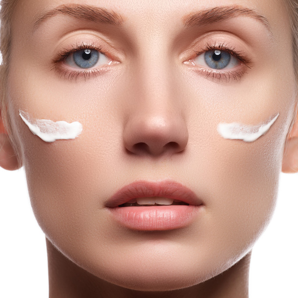 Beautiful face of young woman with cosmetic cream on a cheek. Skin care concept. Closeup portrait isolated on white. Close-up young beautiful face of girl applying moisturize cream. - Valokuva, kuva