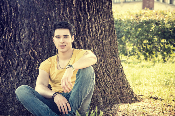 Parkta ağaca yaslanmış çekici bir genç adam. - Fotoğraf, Görsel
