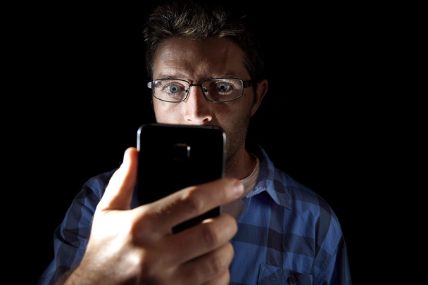 lähikuva nuori mies etsii intensiivisesti matkapuhelimen näytön siniset silmät auki eristetty mustalla taustalla
 - Valokuva, kuva