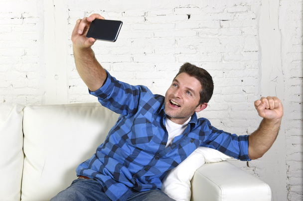 junger attraktiver Mann in den 30er Jahren macht Selfie-Foto oder Selbstvideo mit Handy zu Hause auf der Couch sitzend glücklich lächelnd - Foto, Bild